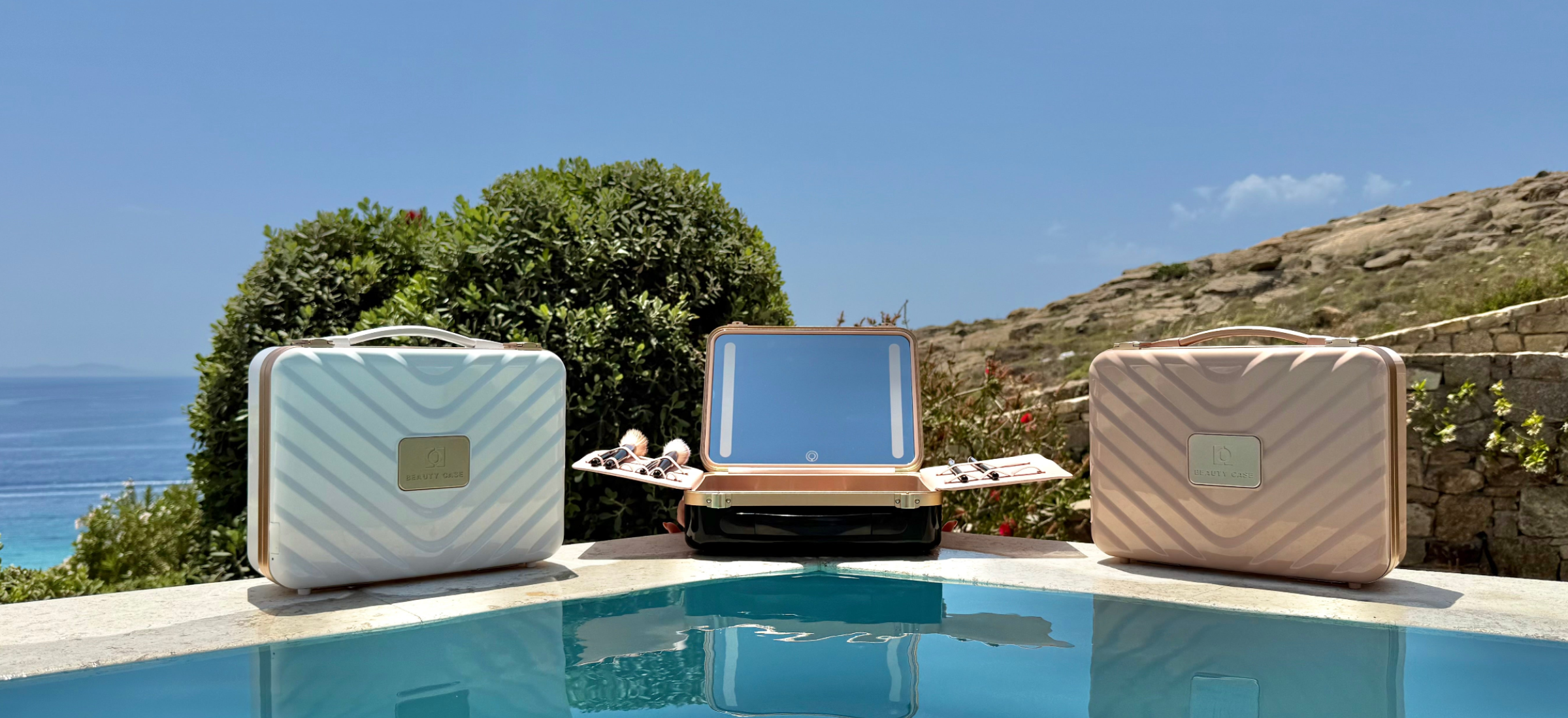 Three Glow & Stow Luxury beauty cases in white, black, and pink, one open showing its mirror - are poolside against a lush green landscape, blue sea, and clear sky on a sunny day.