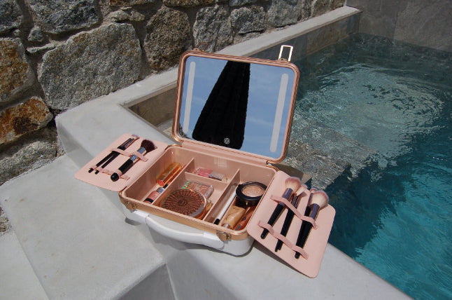 The Glow & Stow Luxury Beauty Case in white, filled with brushes, powders, and cosmetics, sits open by a tranquil swimming pool. Its mirrored lid reflects an object as stone and concrete frame the elegant scene.
